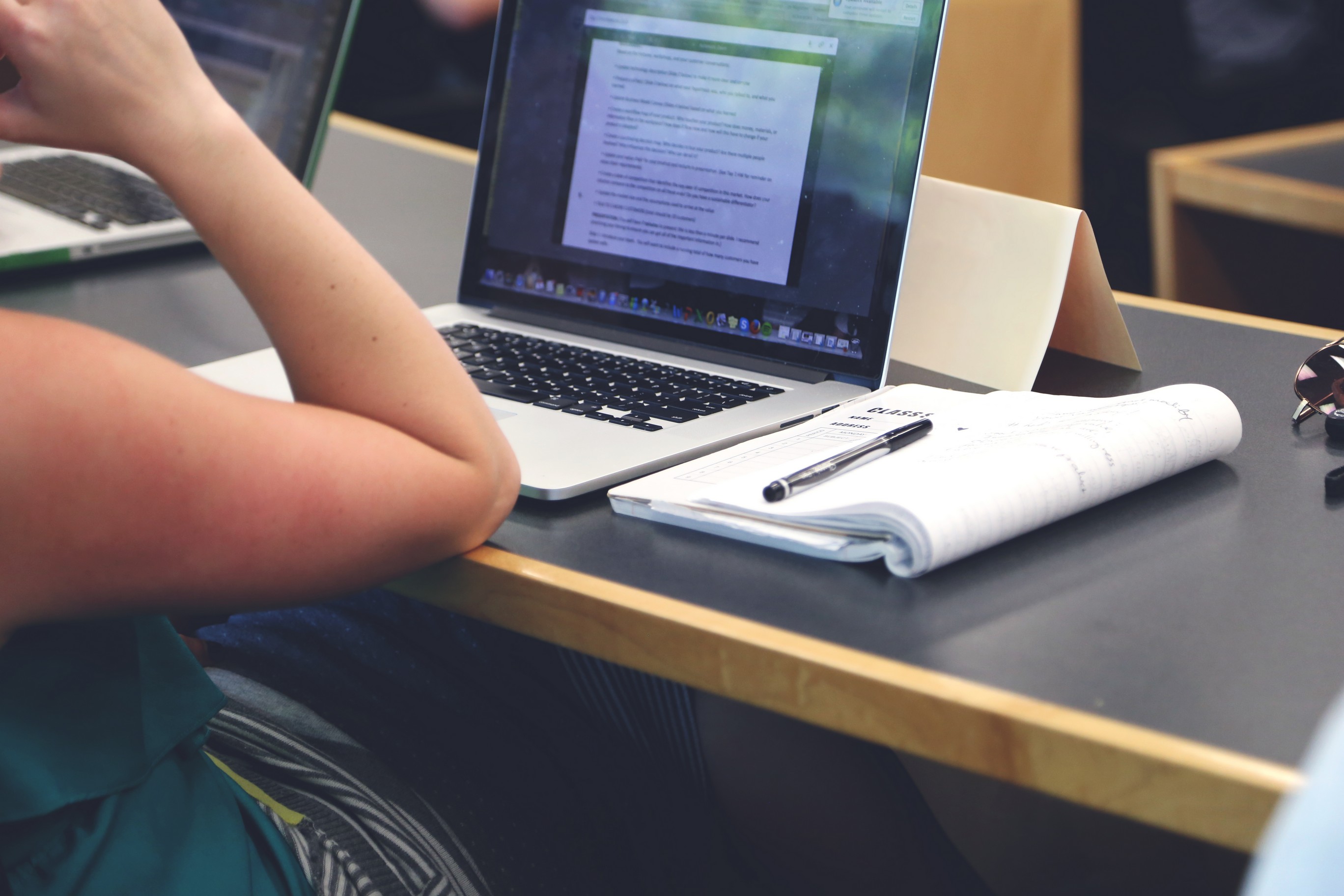 A persons's arm resting on a desk next to a laptop computer and a notebook.