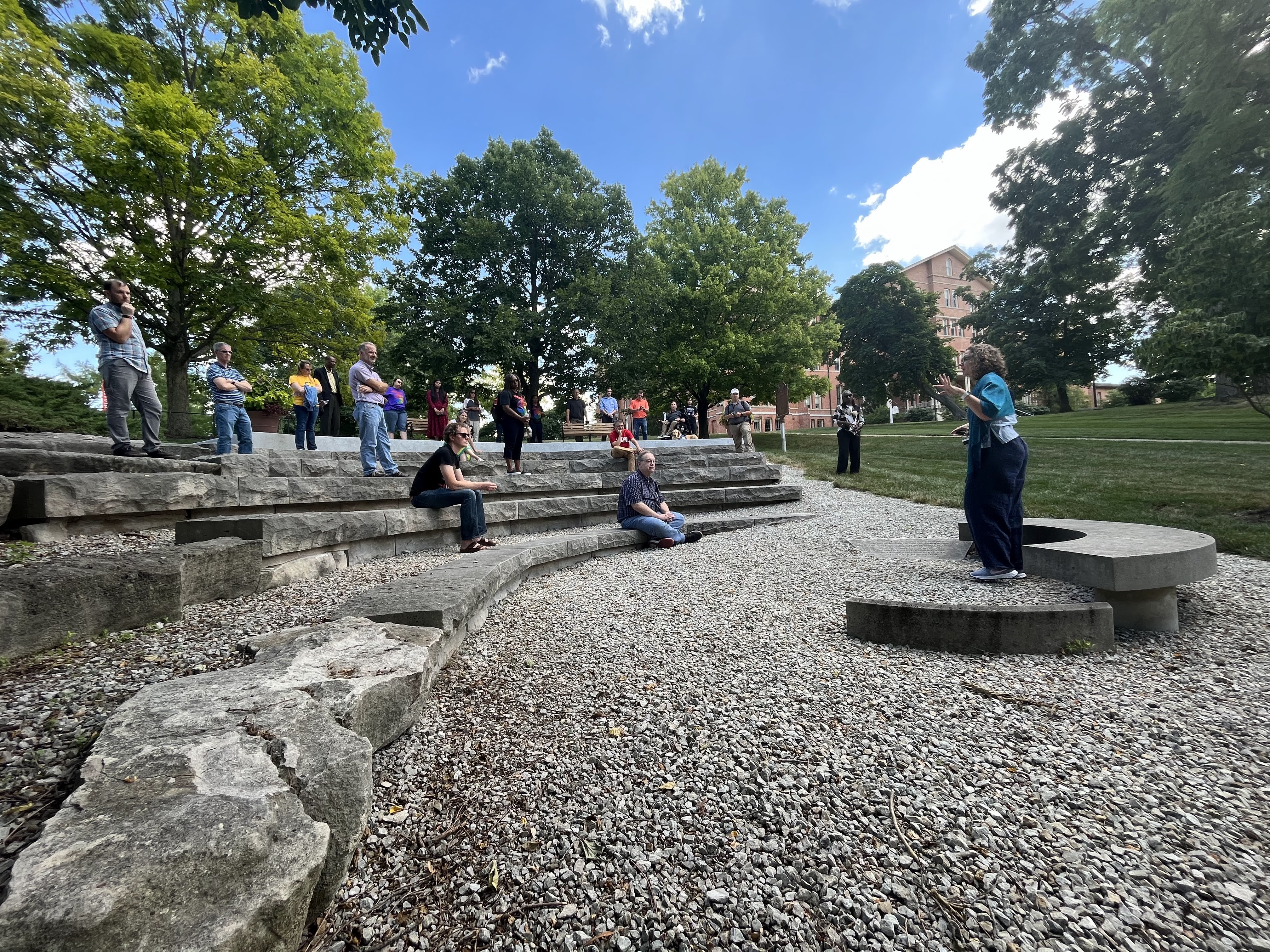 The Freedom Summer memorial seen from the ground up. Members of IT Services sit and stand around the memorial, listening to Ann Elizabeth Armstrong talk about the three young men it was built to memorialize