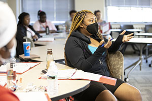 A female-presenting person of color wearing a mask gestures as she speaks