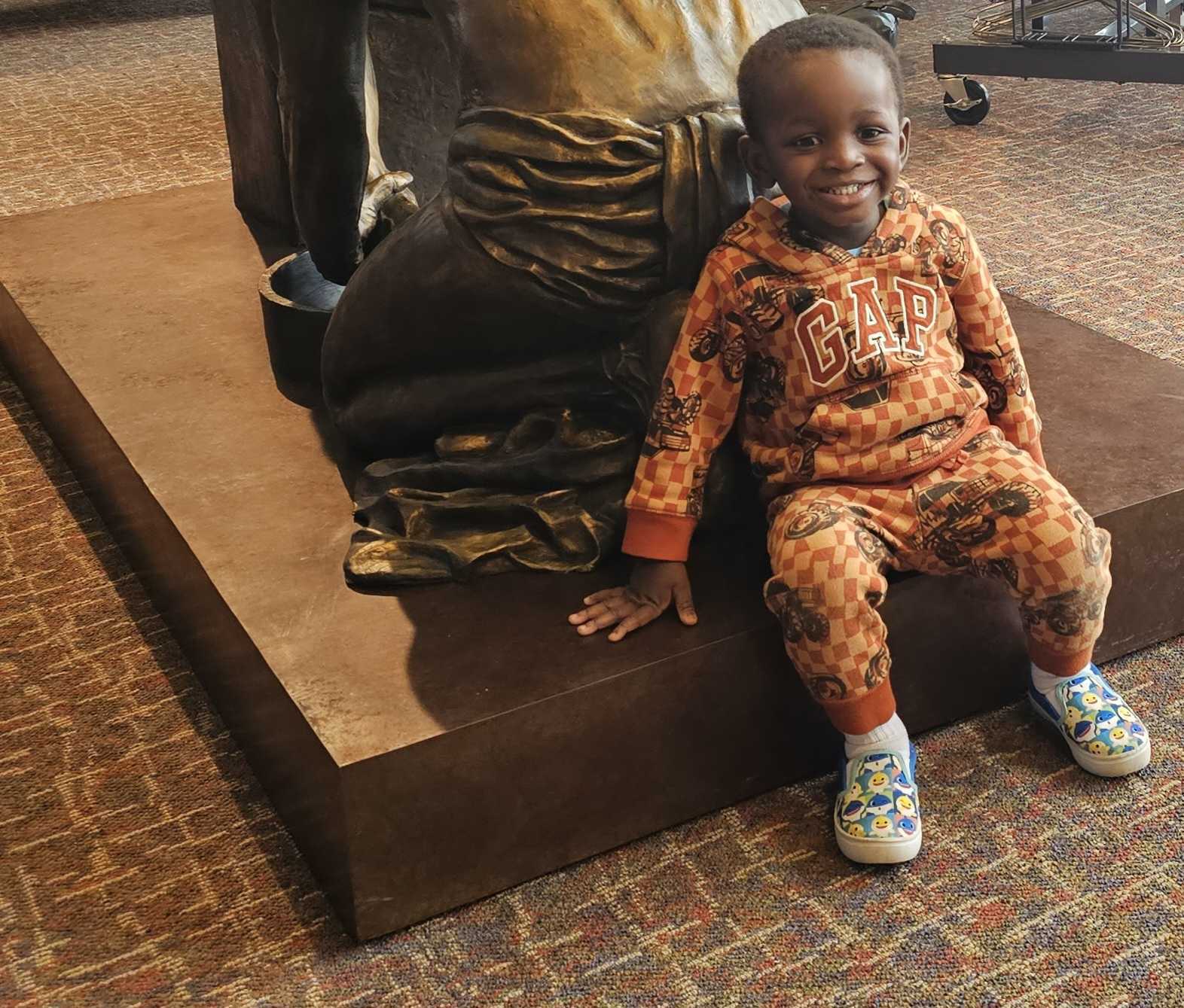 Male presenting child sitting at the base of a statue smiling at the camera. 