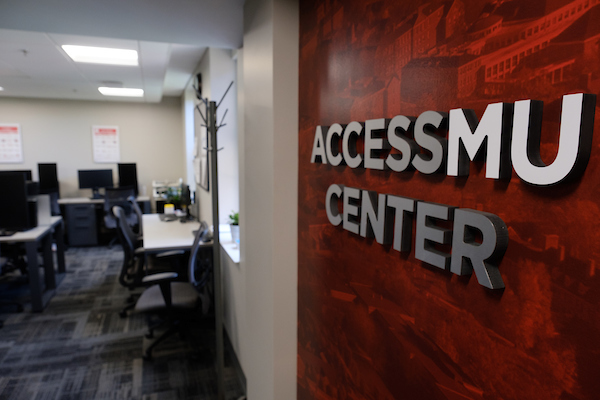 An angled photo of the AccessMU Center with a close up of the wall that reads "AccessMU Center" against a red background. To the left you can see a glimpse of an office space with desks and computers. 