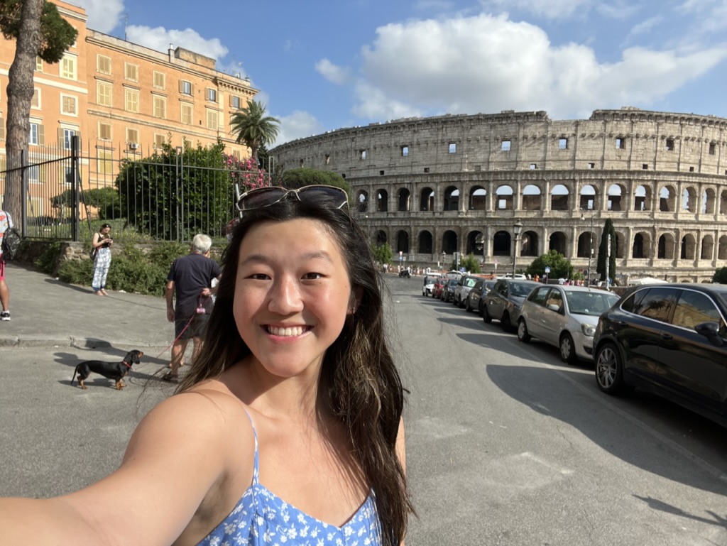Selfie of young asian woman (Serena) smiling at the camera with sunglasses on top of her head and a blue and white sundress. She has long dark hair and brown eyes. In the background is the roman colosseum. The weather is sunny