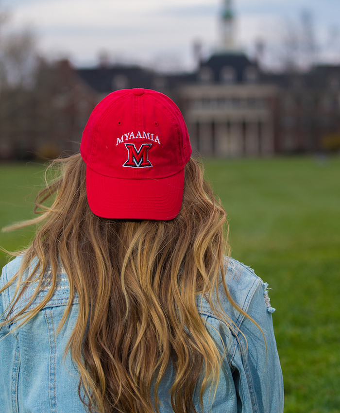 Student sits looking across Central Quad and wearing a red baseball hat. Design includes the Miami University block M logo and the word myaamia above it.