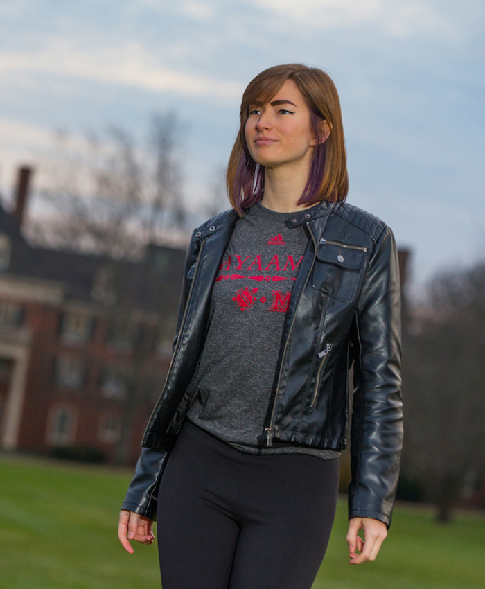 Student wearing a grey t-shirt under a black jacket. The shirt design includes the myaamia heritage logo, Miami University logo, the Adidas logo, and myaamia turtle mark. The word myaamia is above.