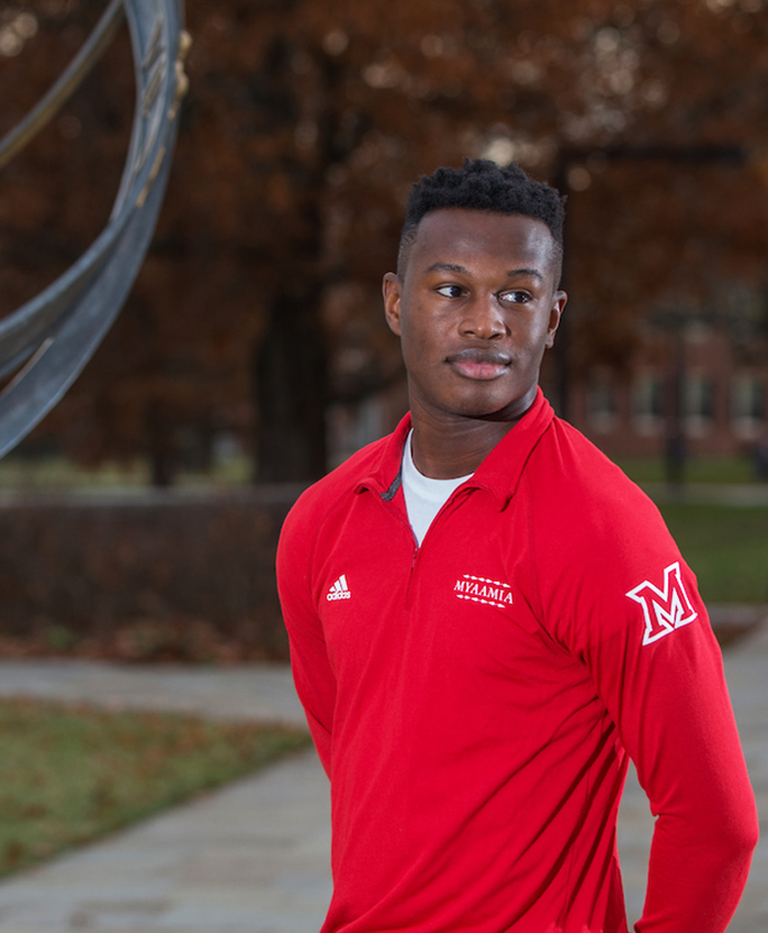 Student stands next to the sundial on central quad. He wears a red quarter zip pullover. The shirt design includes the myaamia heritage logo, Miami University logo, the Adidas logo, and the word myaamia