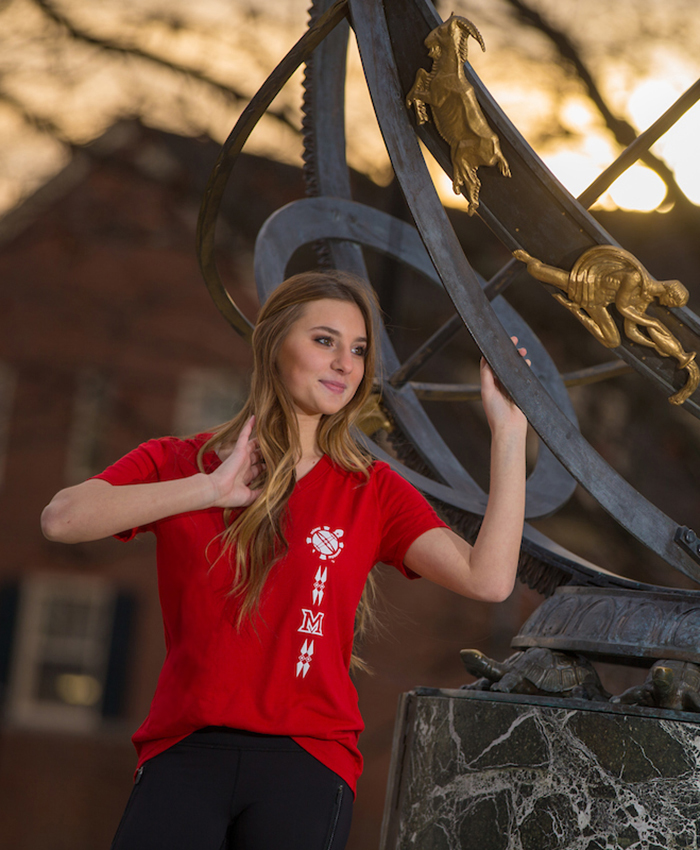 Student stands next to the sundial on central quad. She wears a red t-shirt with a design that includes the myaamia heritage logo, Miami University logo, and the Myaamia Heritage Turtle mark in a vertical pattern