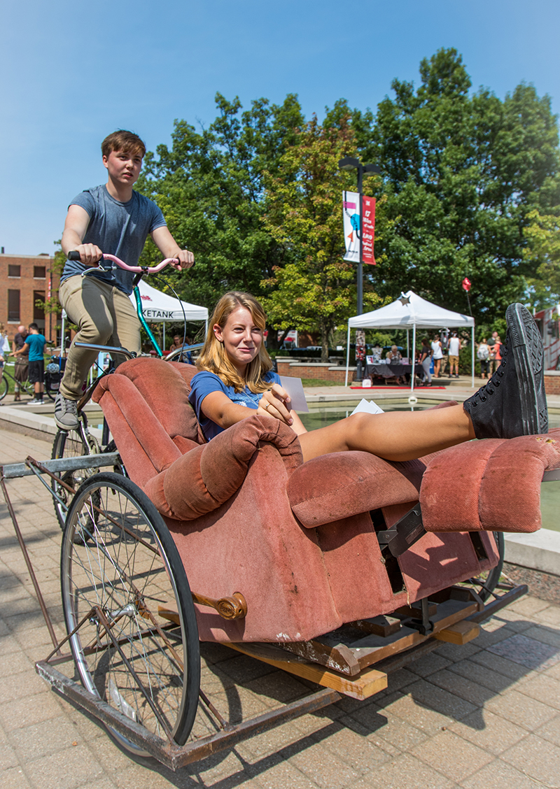 A girl rides in a recliner chair that is attached to a bike driven by a male student