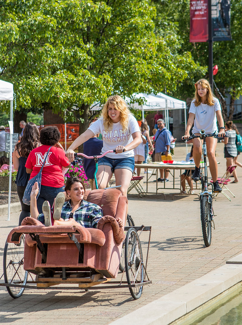 A girl rides in a recliner chair that is attached to a bike driven by a female student while another female student rides a very tall bike in the background