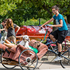 A lady and a dog ride in a recliner chair that is attached to a bike driven by a male student