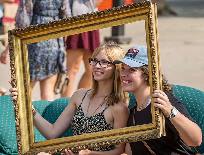 Two female students sitting on a couch posing for a picture while holding up a gold picture frame