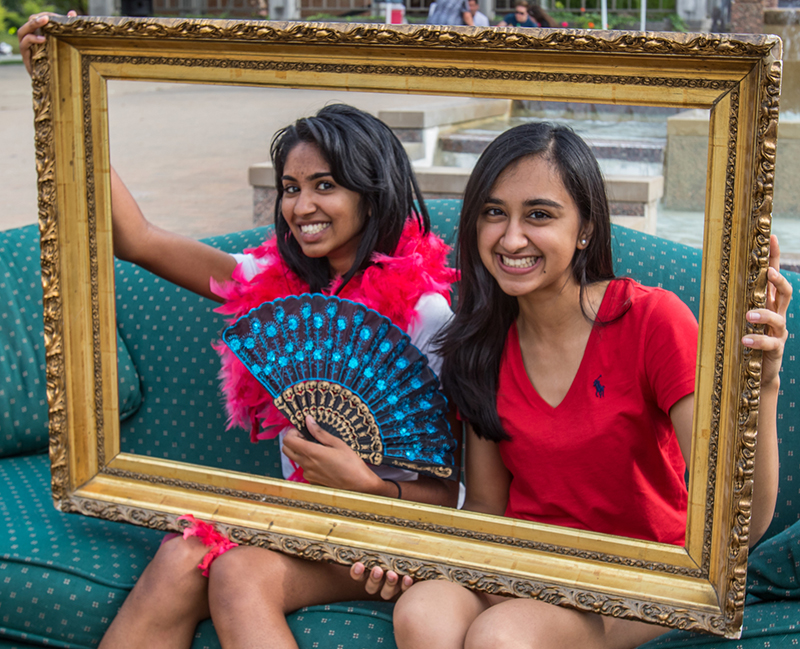 Two girls sit on a couch posing for a picture while holding up a gold picture frame