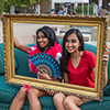 Two girls sit on a couch posing for a picture while holding up a gold picture frame