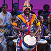 A man dressed in very bright and colorful clothes plays the drums while standing on stage