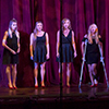 A group of females wearing black dresses and red shoes stands on stage singing