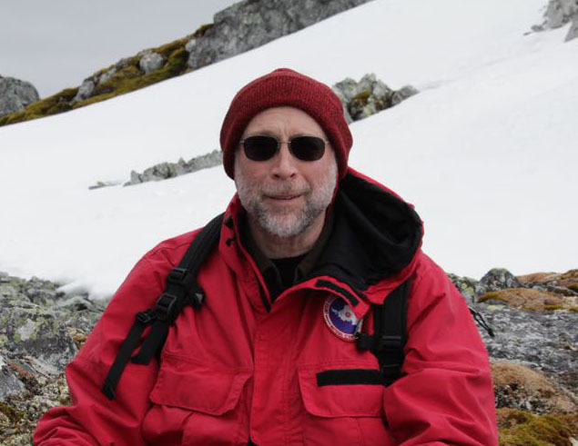 Rick Lee, wearing sunglasses and dressed in winter gear, poses near a snow-covered slope in Antarctica