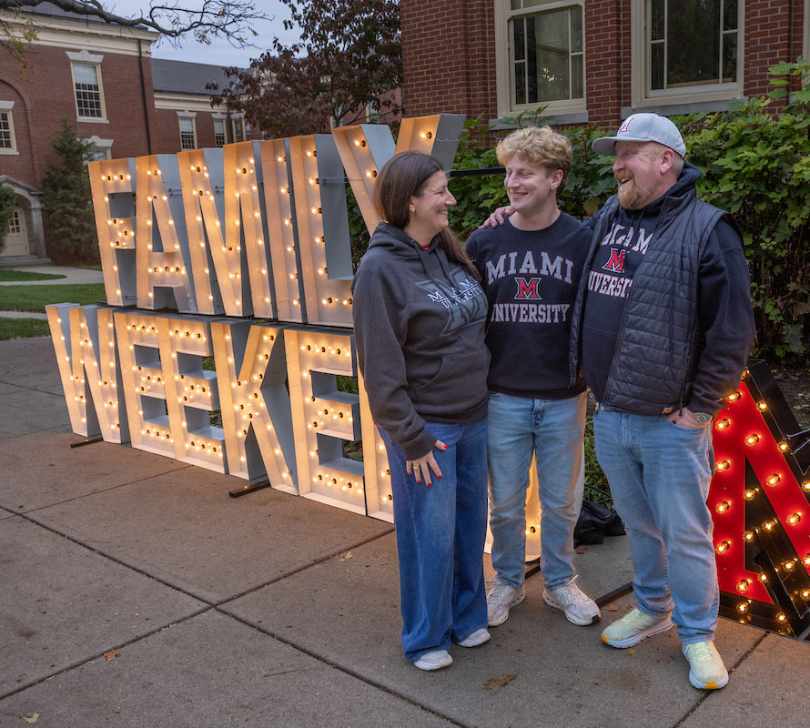 Miami parents pose for a photo op with their student in front of a huge lit up sign that says Family Weekend