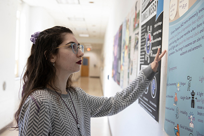A woman in a patterned sweater examines an educational poster on a wall in a well-lit hallway.