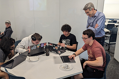 Group of students collaborating at a table, working on laptops while a mentor observes.