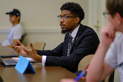 A young man in a suit engages in a discussion at a table during a meeting, with a laptop and notepads in front of him.