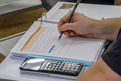 A person writing notes on a paper while holding a pen, with a calculator and documents visible on the table.