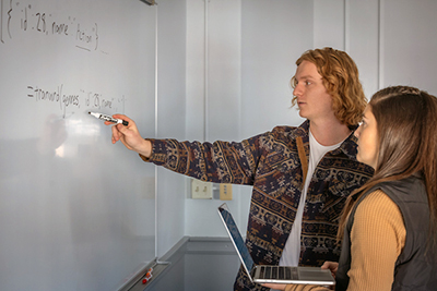 A student explains analytics concepts at a whiteboard to a classmate while holding a laptop.