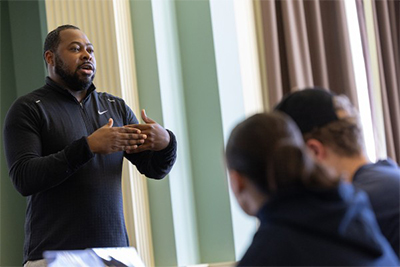A man in a black athletic shirt is gesturing while speaking to a group of students seated at a table.