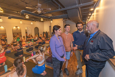 A group of people converses near a yoga class in a spacious studio, with participants practicing yoga on mats in the background.