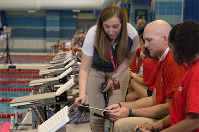 A swim coach instructs a timer at a swimming competition, with competitors in the background ready to race.