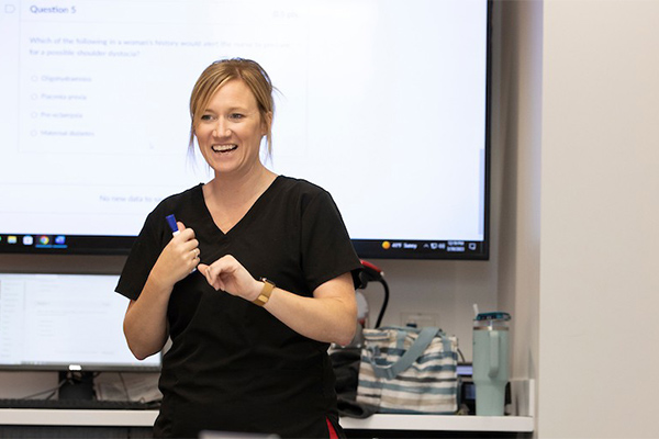 A woman in a black medical scrub enthusiastically teaches a class, with a digital screen displaying a question in the background.