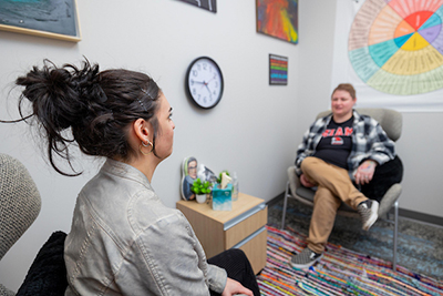 A woman sits attentively in a therapy session with another individual, surrounded by colorful artwork and a clock on the wall.