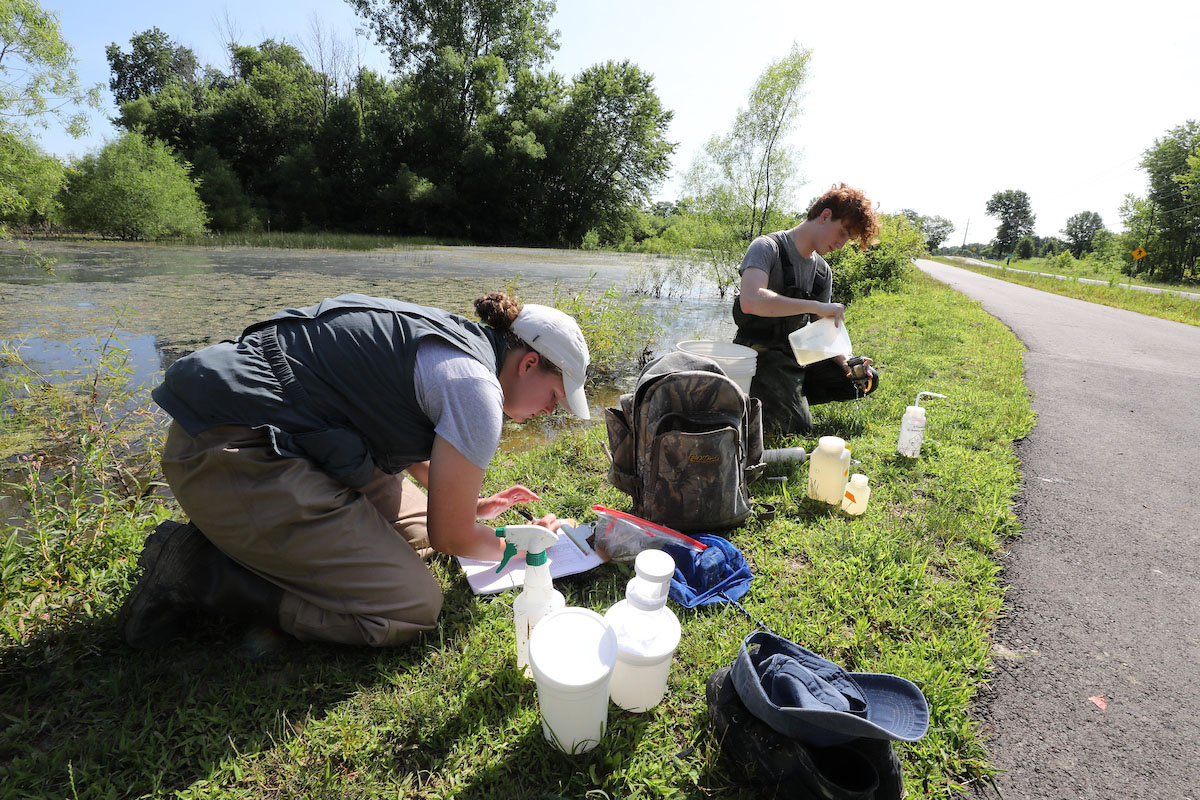 Two ecology students taking notes and collecting samples by the water