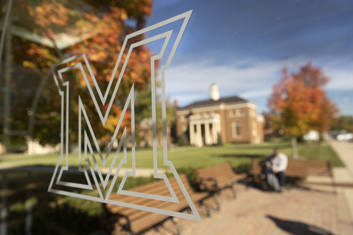 The Miami logo etched on glass with the scenic Oxford campus reflecting off of the glass