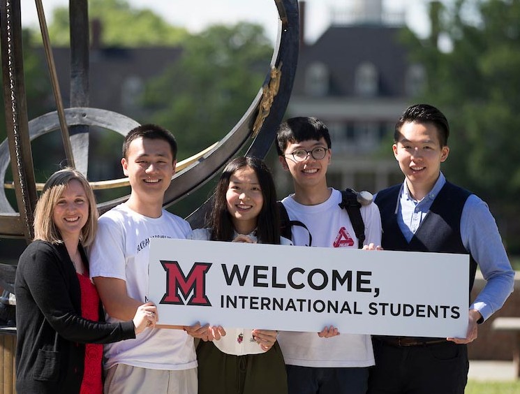 Un grupo de estudiantes internacionales se para frente al reloj solar de Miami sosteniendo un cartel de bienvenida a los nuevos estudiantes internacionales.