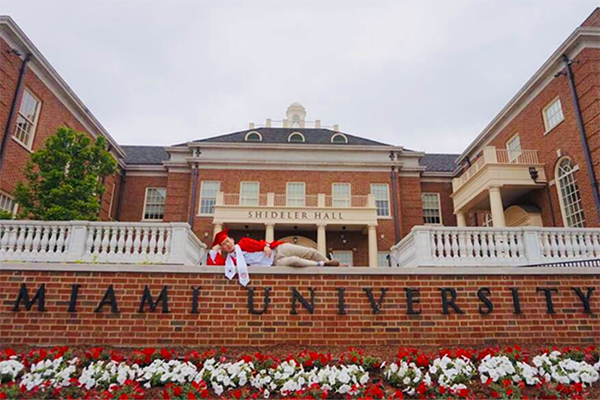 James Heo posing in front of Shideler hall wearing his graduation cap and gown