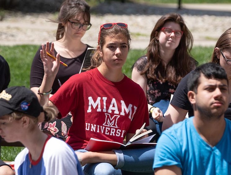 Una estudiante levanta la mano durante una clase al aire libre en un día soleado.