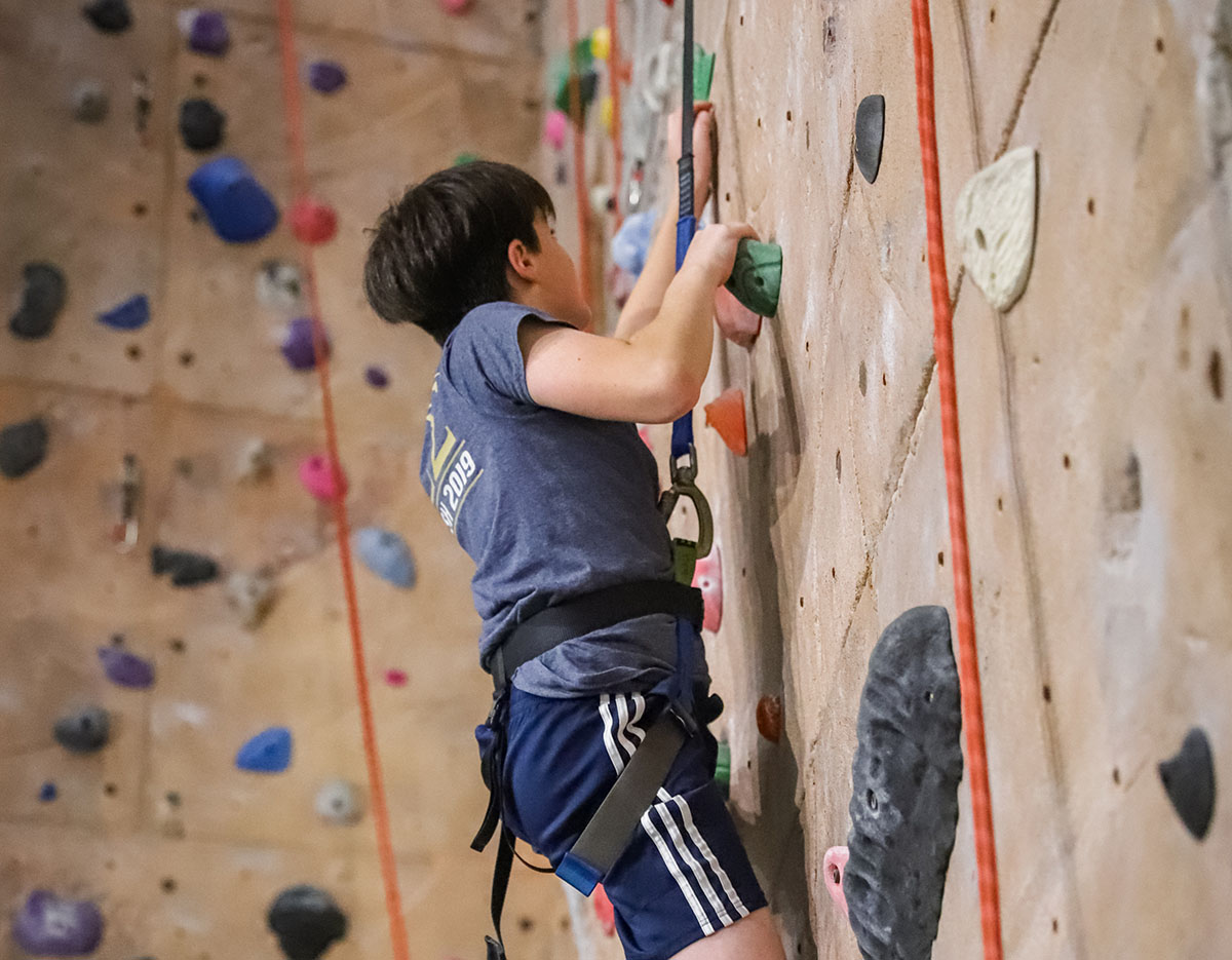 Young boy climing the rock wall wearing a harness with ropes