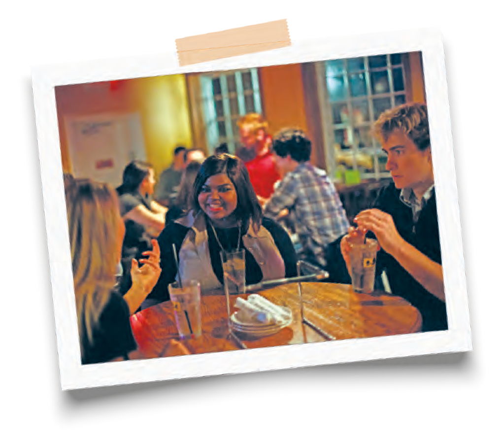 Three people sit at a round café table with drinks, chatting while other patrons blur in the background.