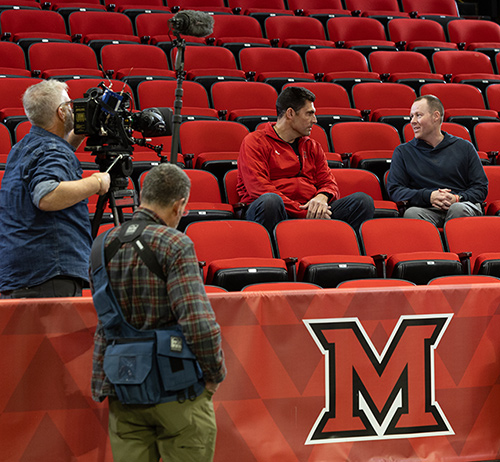 Two men talk while seated among rows of red arena seats as a camera crew films nearby, with a large letter M banner in the foreground.
