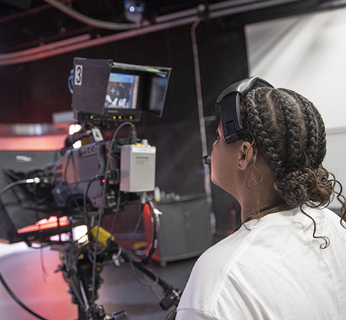 Person with braided hair wearing a headset operates a studio camera in a TV production set.