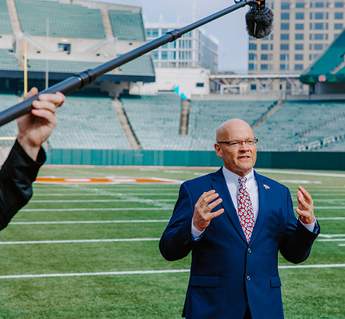 Man in a suit speaks on a football field while a boom microphone is held overhead, with stadium seats in the background.