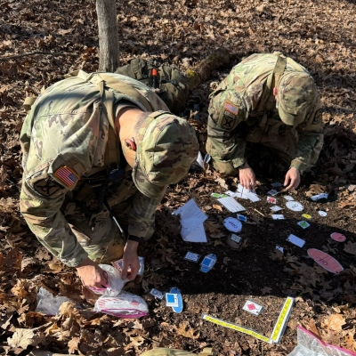 Two soldiers in camouflage kneel in a wooded area, arranging training markers and notes on the ground.