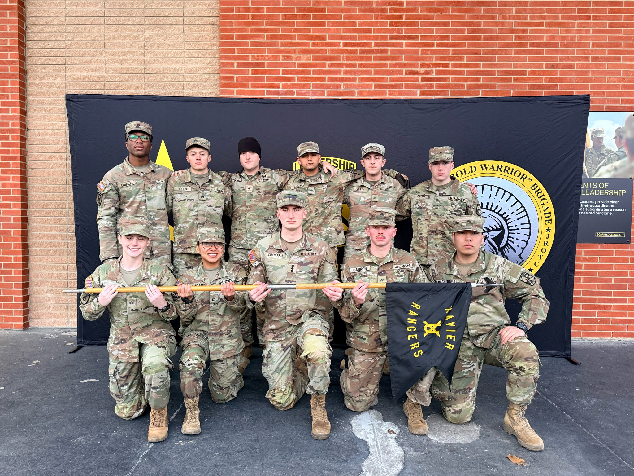 Army ROTC cadets in camouflage uniforms pose in front of a unit banner, holding a guidon that reads “Xavier Rangers.”