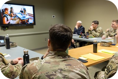 Soldiers in uniform sit around a conference table watching a presentation on a wall-mounted TV in a meeting room.