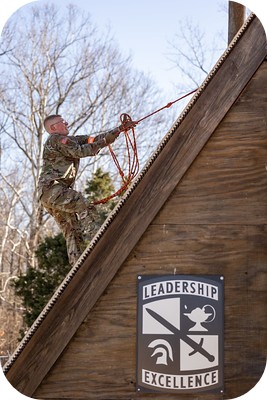 Person in camouflage climbing a steep roof using a rope next to a “Leadership Excellence” sign.