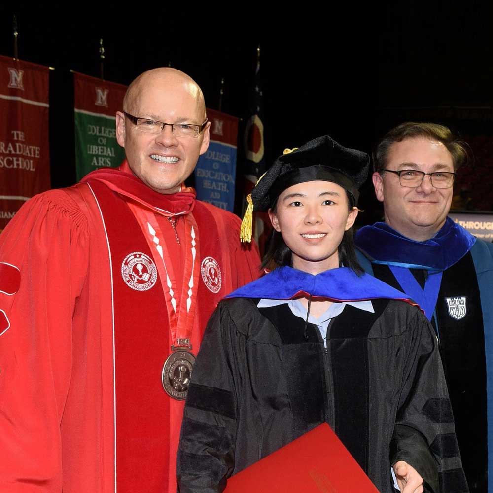 Miami University President Greg Crawford with recent doctoral graduate Xiao Qiu and J. Scott Brown, Chair of the Department of Sociology and Gerontology, all dress in graduation robes and caps.