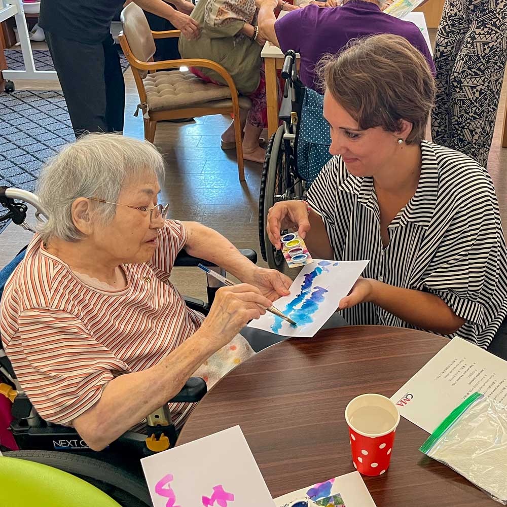 A young woman kneels next to an older Japanese woman while they both hold paper and blue paints. 