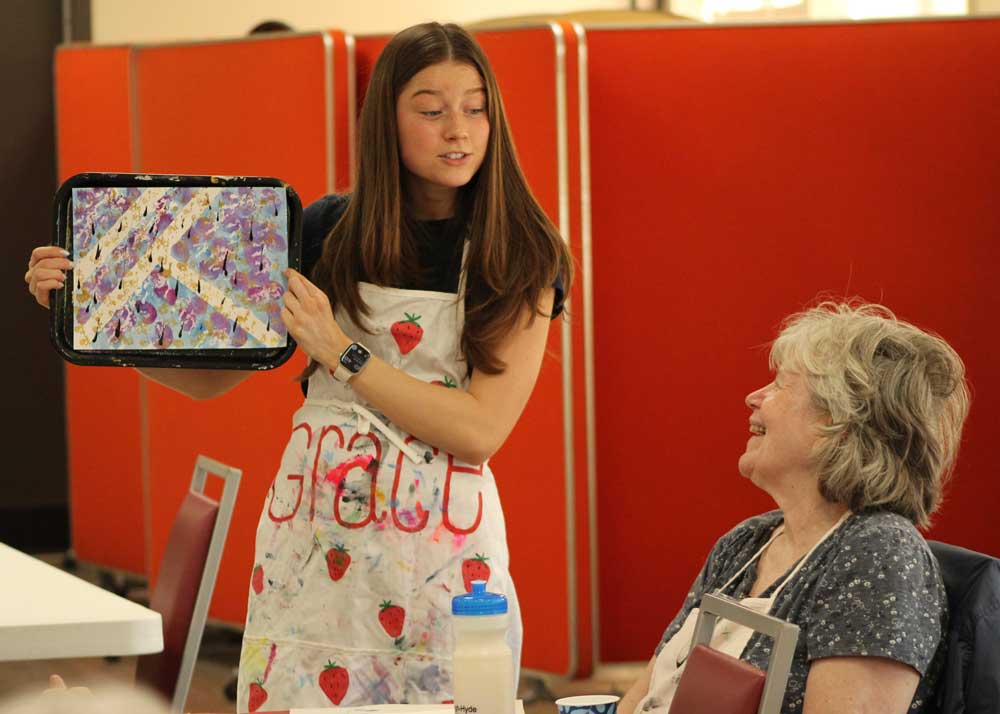 A young student holding up OMA artwork with an older woman in a wheelchair smiling. 