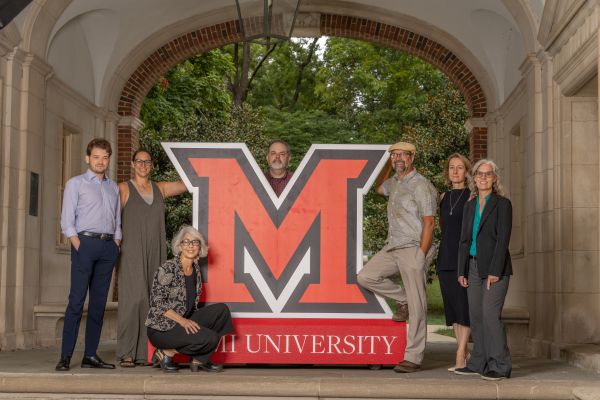 The anthropology department faculty. From left to right, Professor Rhue in a light blue shirt and dark blue pants. Dr. Ellis in a green dress with glasses, Dr. Hay-Rollins kneeling with a black dress and floral patterned coat. Dr. Card with a red shirt and a beard, Dr. Mark Peterson wears beige pants and a grey button up shirt with a tan hat. Dr. Klumbyte wears a black dress with a silver necklace, Mrs. Keener wears a teal shirt with a black coat and dark grey pants.