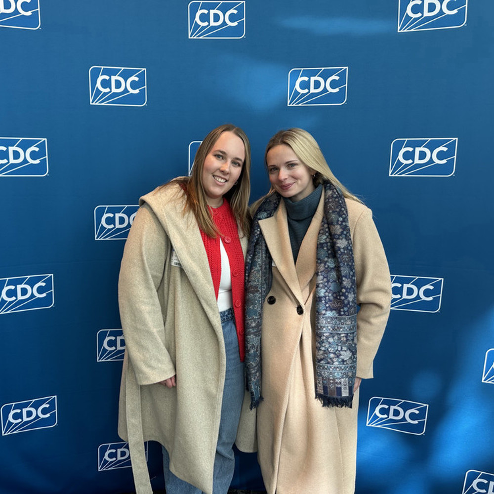 Two women pose for a picture in front of a blue banner with the Centers for Disease Control logo.