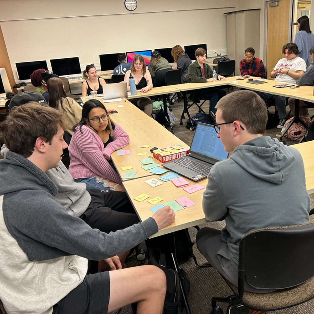A group of surround a computer and talk while they write on post-it notes.